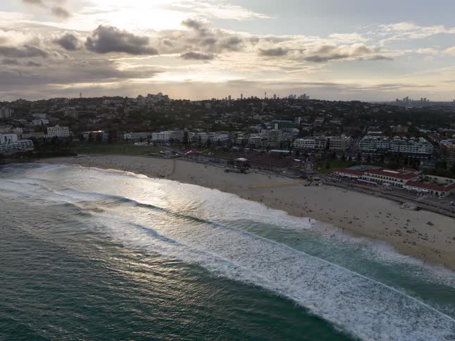 An epic aerial view flies over the ocean towards a massive festival on a crowded beach at sunset with a city skyline in the distance
