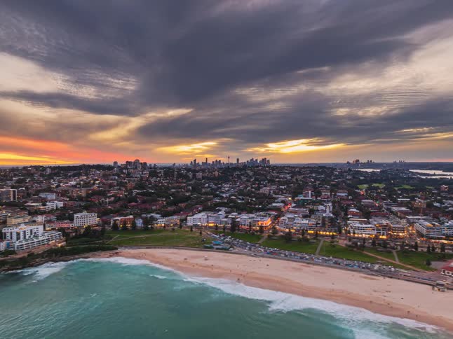 A captivating aerial time-lapse shows the transition from a moody sunset to the illuminated city lights over Bondi Beach and the Sydney skyline