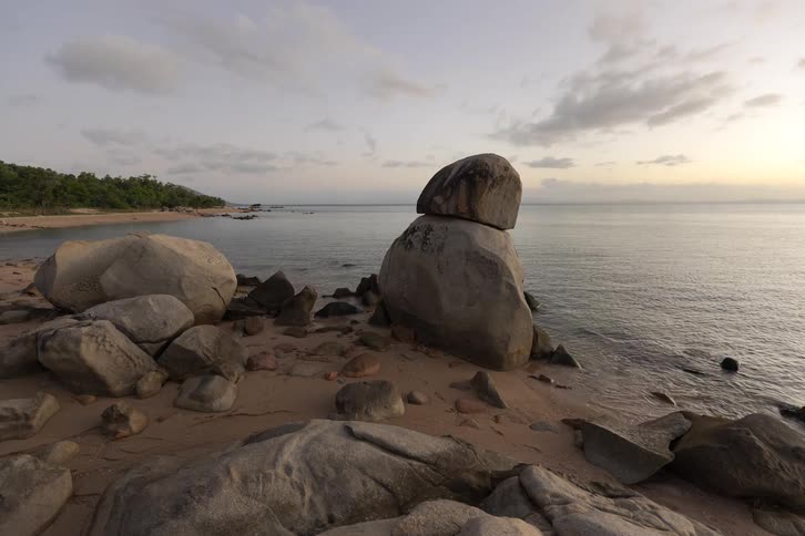A time-lapse captures a peaceful sunset over a rocky coastline with a unique balancing boulder formation