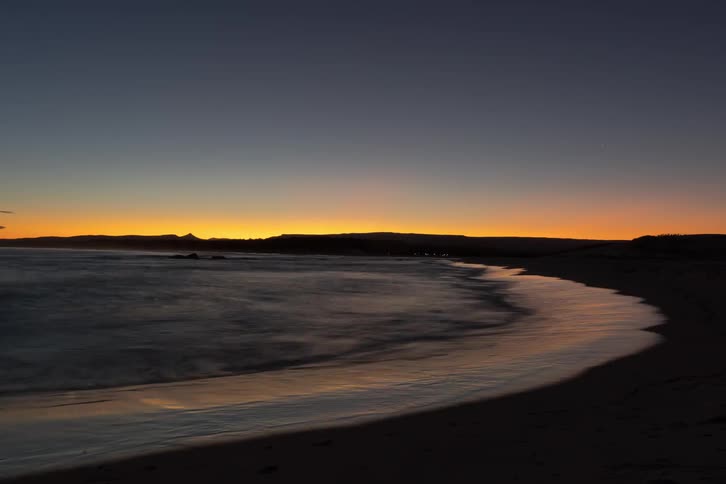 A tranquil time-lapse captures the gentle motion of waves on a sandy beach as the last light of sunset glows on the horizon