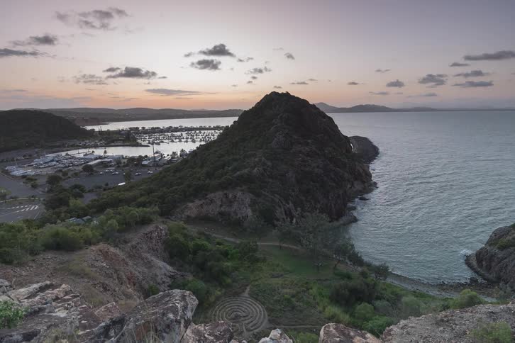 A scenic day-to-night timelapse shows a colorful sunset over a coastal harbor and a large, rocky hill