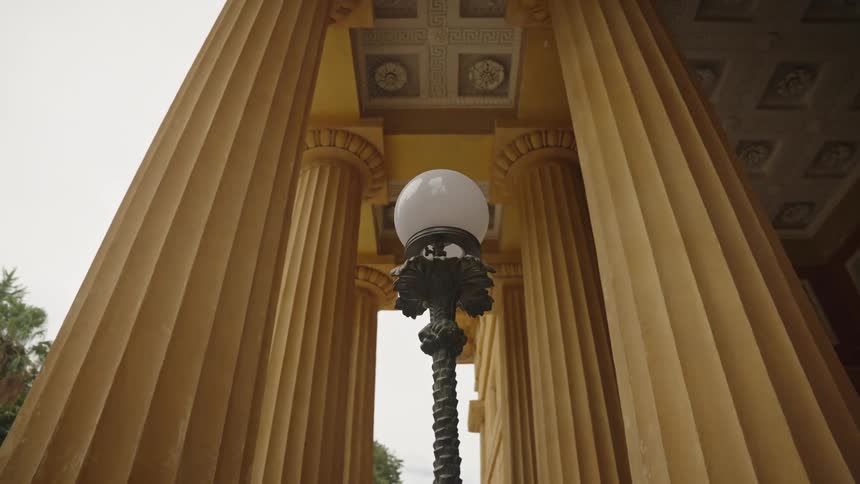 An ornate vintage street lamp stands before the majestic yellow columns of a classical building in a slow pull-out shot