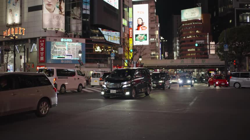 Cars drive through a bustling intersection in Shibuya, Tokyo, illuminated by glowing billboards and city lights at night