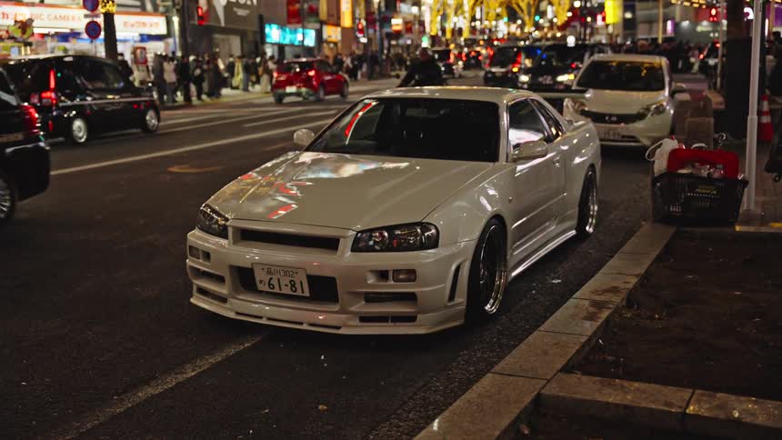A sleek white sports car is parked on the side of a bustling, brightly lit street in Tokyo at night as traffic passes by