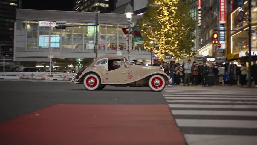 A vintage tan convertible drives slowly through the bustling, brightly lit streets of Shibuya, Tokyo, at night as crowds watch from the sidewalk