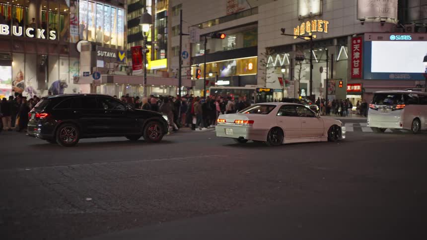 Cars and taxis navigate the iconic Shibuya Crossing in Tokyo at night, surrounded by bright neon signs and crowds of people