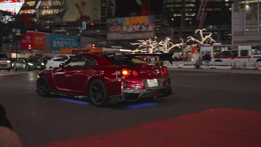 A modified red sports car with bright blue underglow lights slowly navigates a busy, neon-lit intersection in Tokyo at night