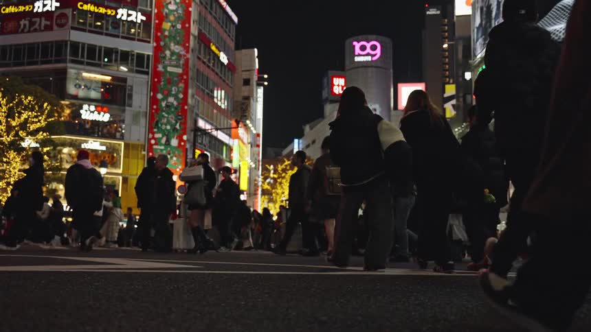 A low-angle, slow-motion view captures the endless stream of pedestrians crossing the iconic Shibuya Scramble at night in Tokyo