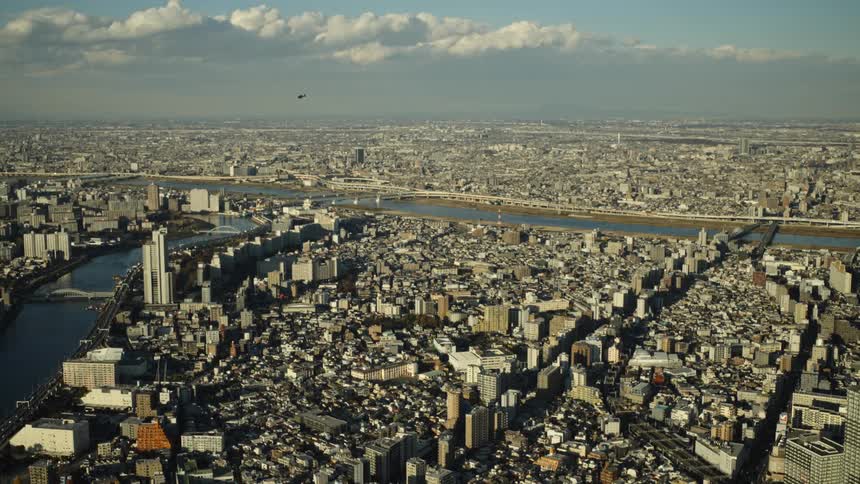 A wide aerial shot shows the vast, sprawling cityscape of Tokyo with rivers cutting through the dense urban landscape as a helicopter flies by