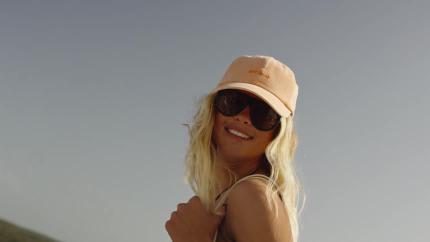 Slow motion, low-angle close-up of a smiling blonde woman in a cap and sunglasses on a sunny day