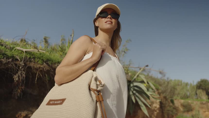 A stylish woman in sunglasses and a cap walks along a sunny beach cliffside with her tote bag in slow motion