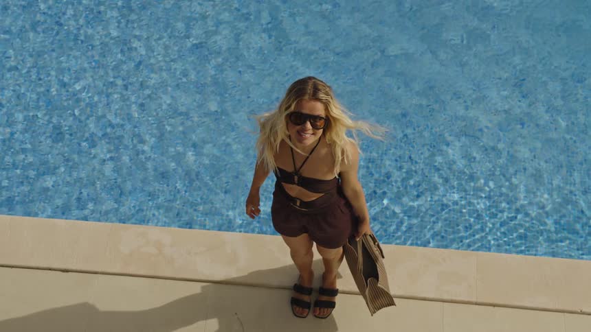 High angle slow motion shot of a stylish blonde woman smiling while standing by the edge of a bright blue swimming pool
