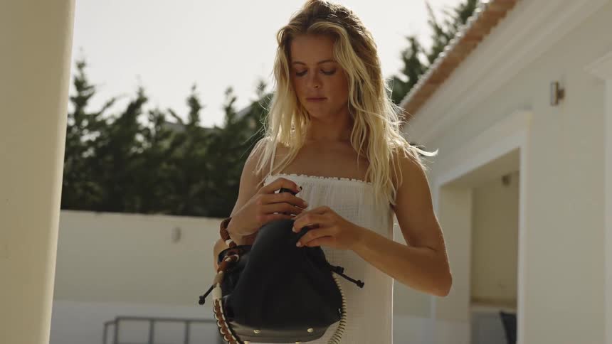 A beautiful blonde woman in a white summer dress cleans her sunglasses before putting them away in her stylish bag