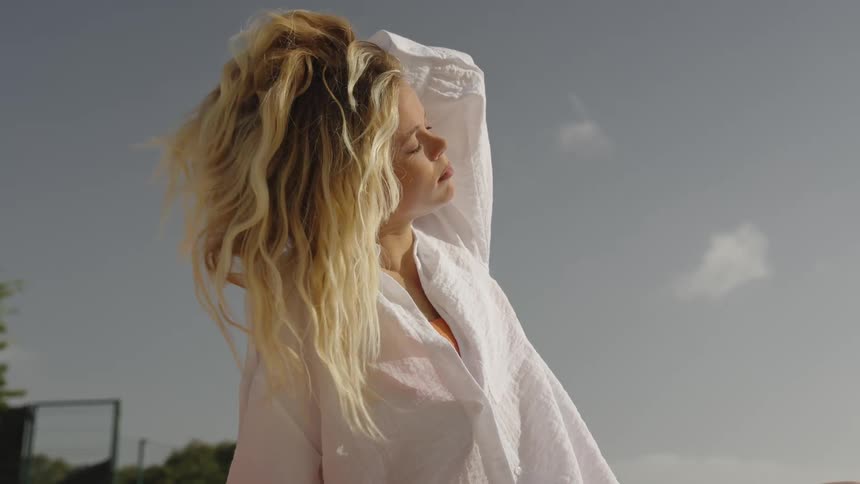 A serene blonde woman in a white shirt enjoys the warm sun as the wind blows through her hair in slow motion