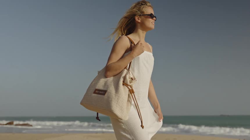 A beautiful blonde woman in a white dress and sunglasses walks along a sandy beach in slow motion with her hair blowing in the wind