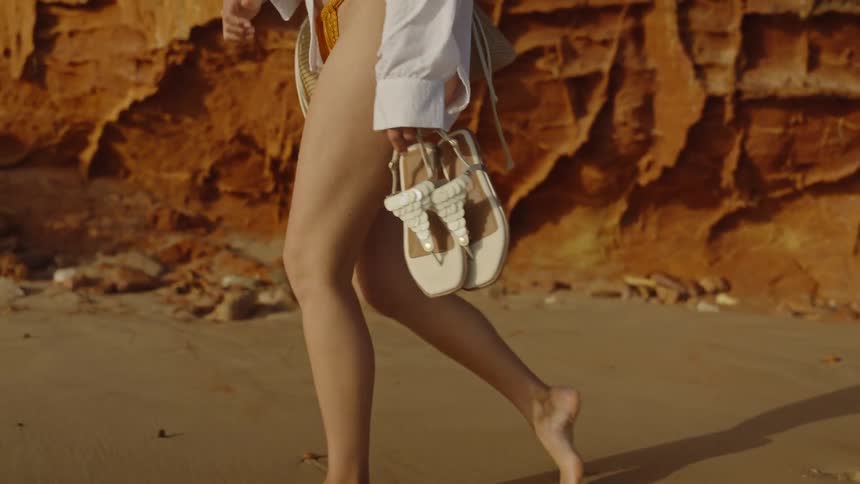 A woman in a bikini and white shirt walks barefoot along a sandy beach next to red cliffs, carrying her sandals and a woven bag