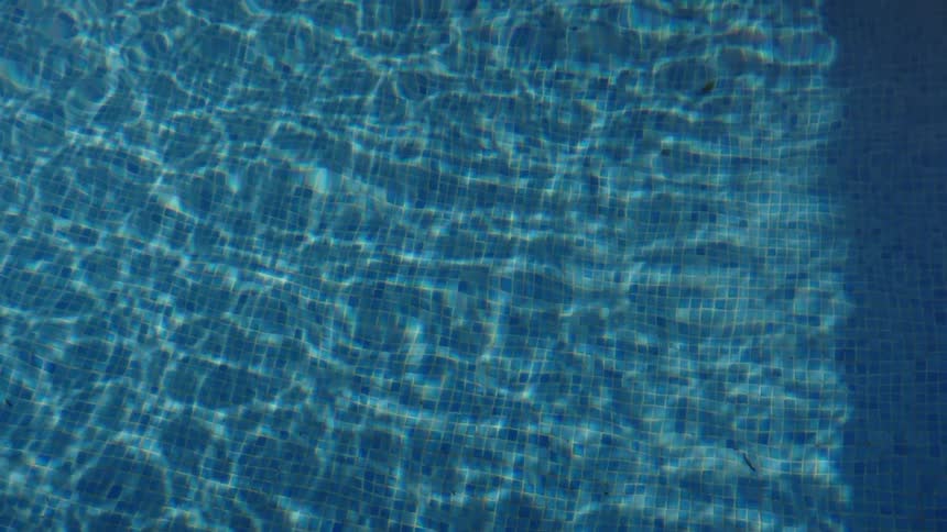 An overhead shot of crystal clear water rippling in a blue tiled swimming pool, with sunlight creating mesmerizing reflections