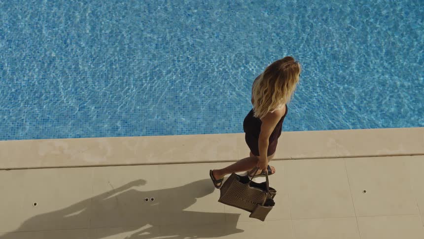 High angle shot of a beautiful blonde woman in a stylish brown outfit walking by a luxurious swimming pool