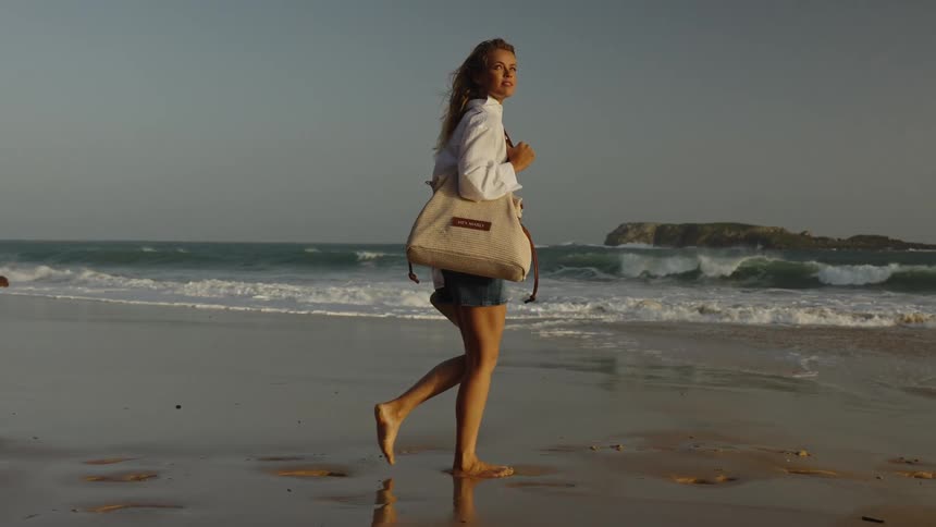 A stylish woman enjoys a serene walk along the shore at golden hour, unbuttoning her shirt as the ocean waves crash behind her