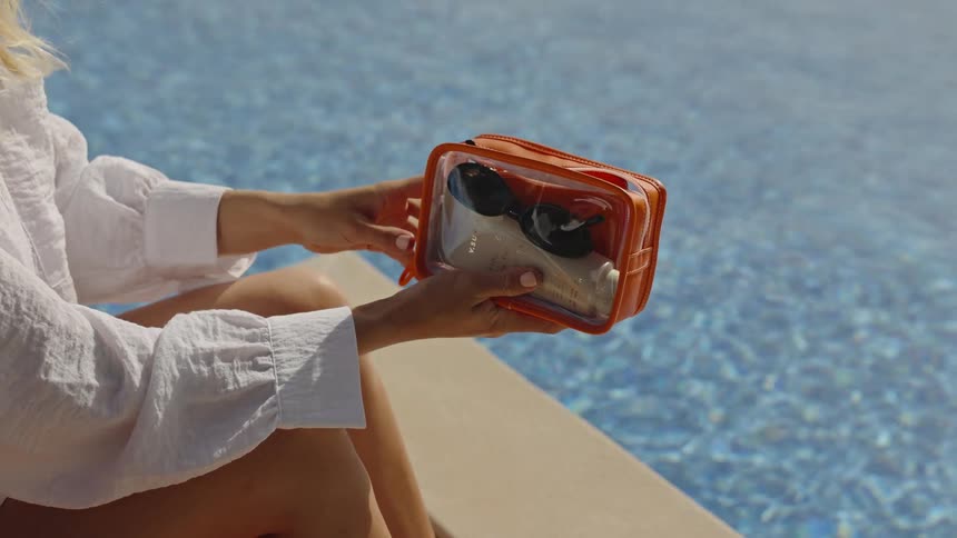 A woman's hands carefully place a pair of sunglasses into a clear and orange cosmetic bag and zip it closed while relaxing by a sunny swimming pool