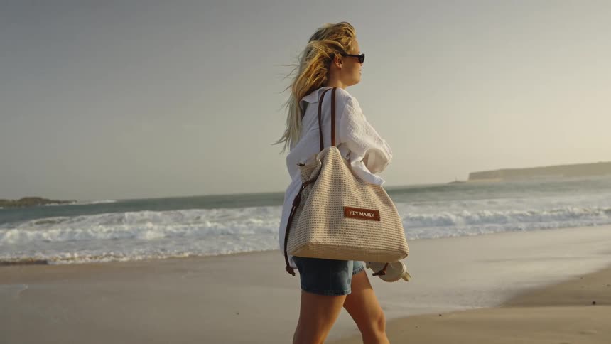 A stylish blonde woman carrying a tote bag walks along the sandy shoreline in slow motion as the ocean waves crash behind her