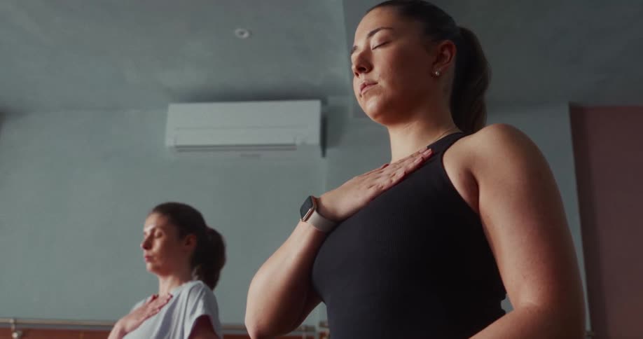 Two women practice mindfulness and deep breathing meditation with their hands on their chests in a calm studio
