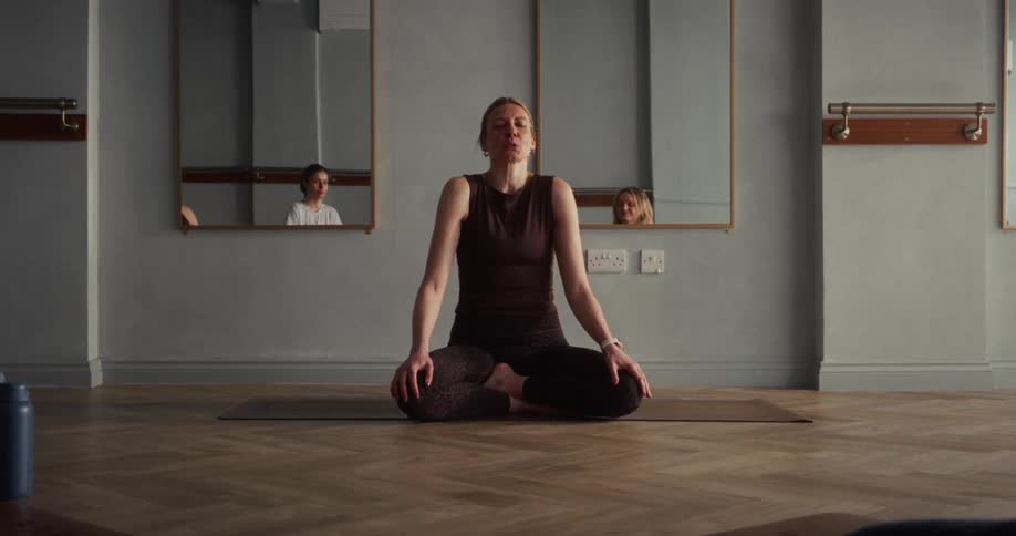 A calm yoga instructor leads her class into a meditative breathing exercise while sitting cross-legged in a serene studio