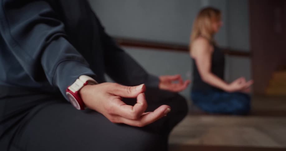 A close-up of a woman's hand in a Gyan mudra pose while meditating with another person in a yoga studio