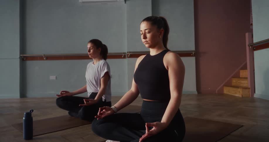 Two women with their eyes closed peacefully meditate in a lotus position during a yoga class