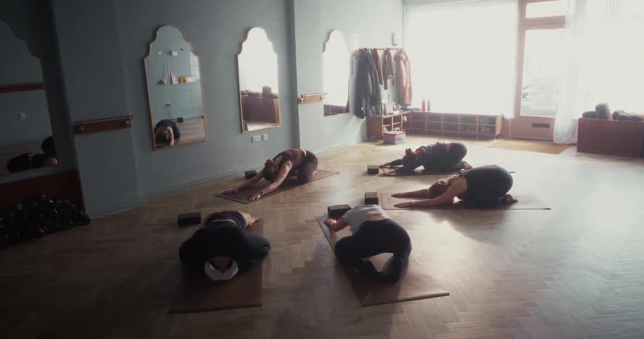 A group of women hold a relaxing child's pose during a yoga class in a sunlit studio
