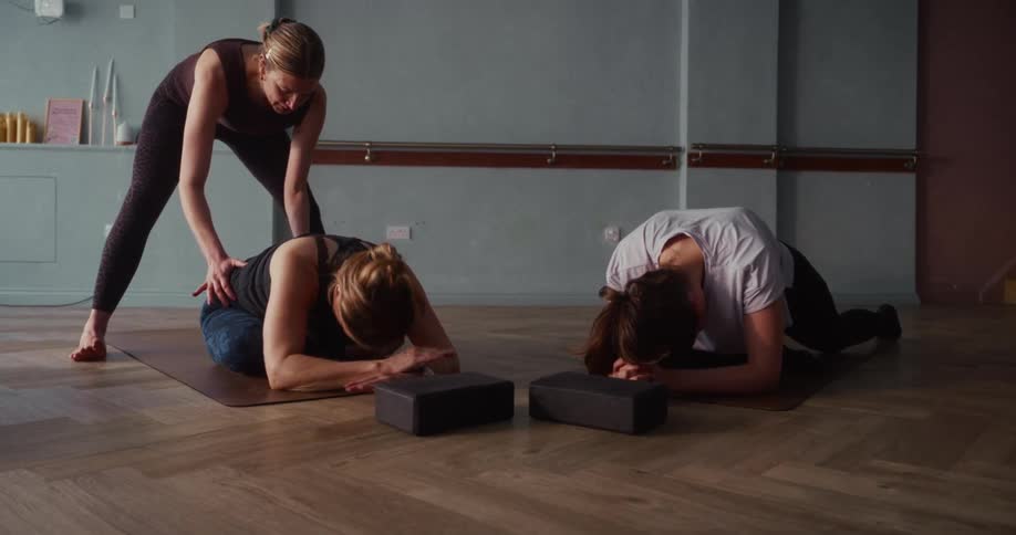 A yoga instructor assists a student with her form during a deep stretching pose in a calm studio