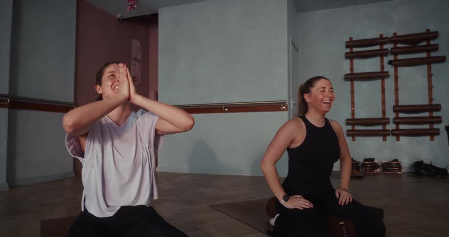 Two happy female friends laugh and smile while practicing yoga together in a modern fitness studio