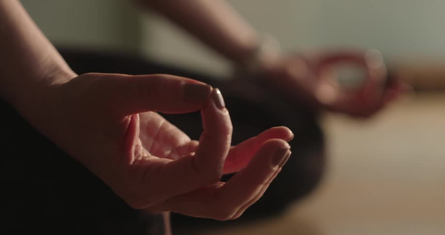 A close-up shot focuses on a woman's hand in a mudra pose while she meditates in a tranquil, softly lit room