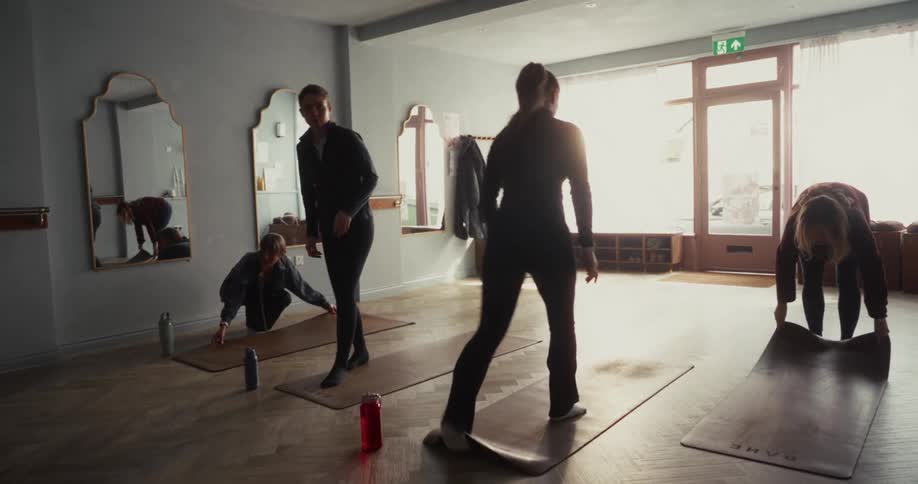 Women unroll their yoga mats on the wooden floor of a sunlit studio, preparing for a class to begin