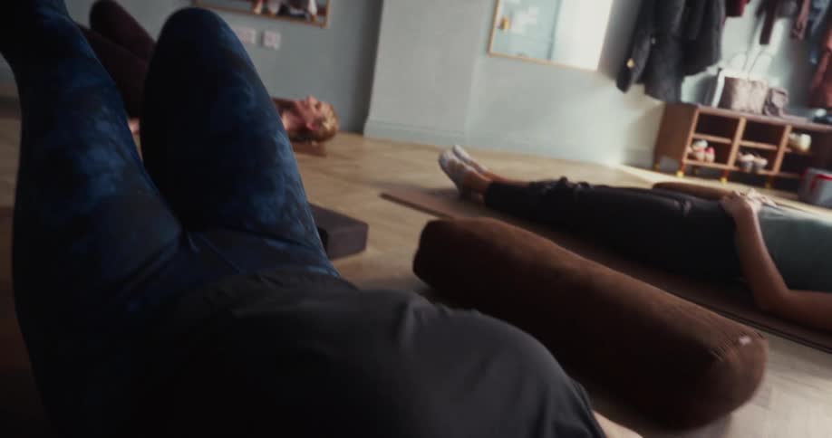 Low angle camera tilt down from a woman's bare foot to her face as she lies in Shavasana pose during a yoga class