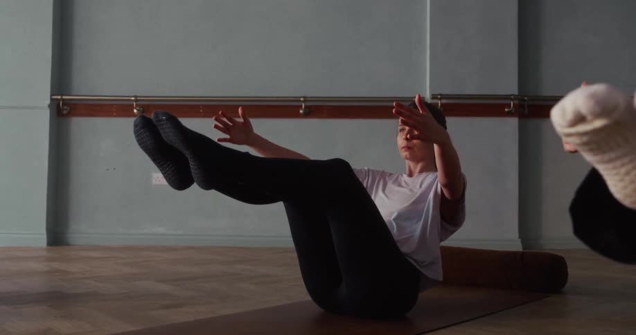 A woman in a sunlit studio holds a challenging boat pose on a yoga mat during a fitness class, filmed in slow motion