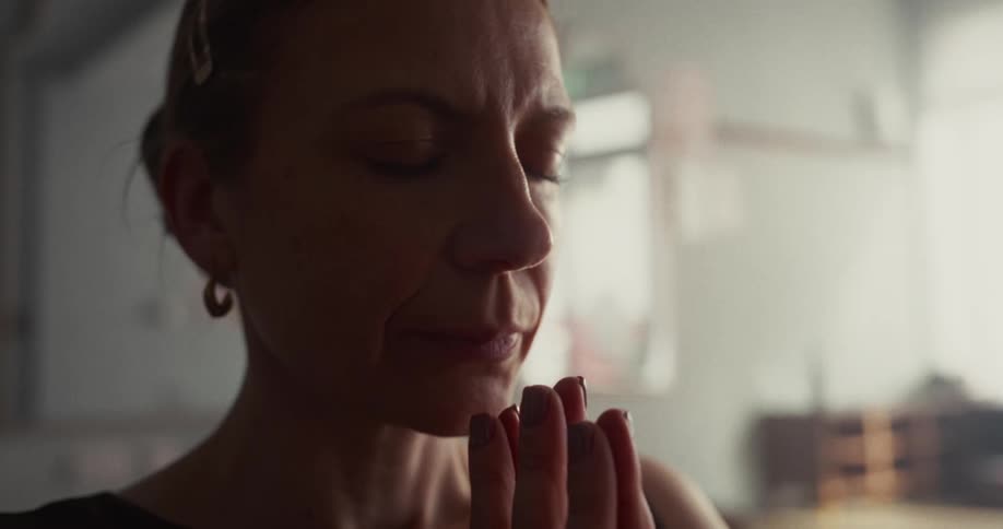 A close-up of a serene woman meditating with her hands in a prayer position in a softly lit room