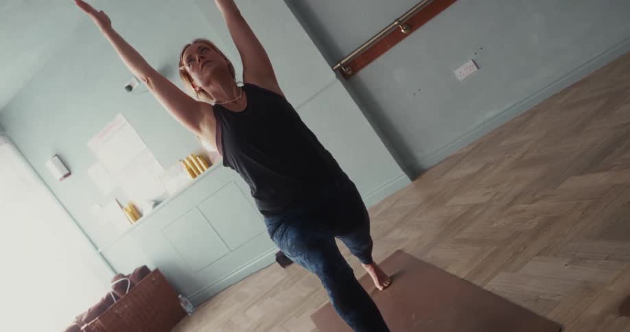 A focused woman transitions from a balancing yoga pose to a high lunge in a beautiful, calm studio in slow motion