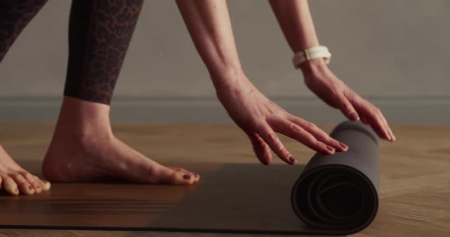 A barefoot woman rolls up her black yoga mat on a wooden floor in slow motion after her workout