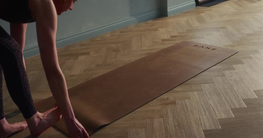 A barefoot woman in athletic leggings slowly unrolls her yoga mat on a herringbone wood floor in a studio