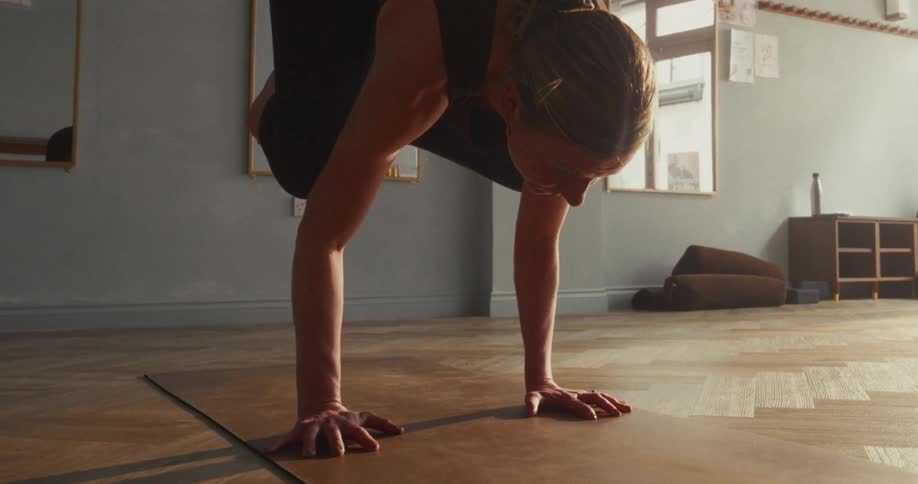 A focused woman holds a challenging arm-balancing yoga pose on her mat in a sunlit studio