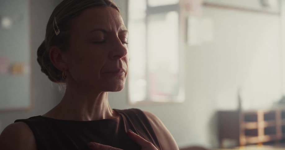 A serene woman meditates with her eyes closed and a hand over her heart in a softly lit room