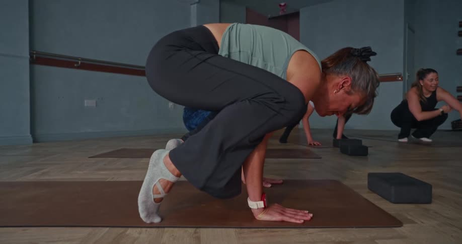 A focused woman in a yoga class attempts a challenging crow pose in slow motion