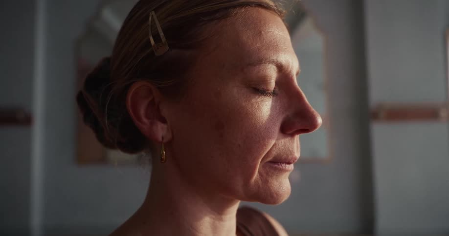 A serene middle-aged woman's face is illuminated by soft light as she meditates with her eyes closed in a quiet room