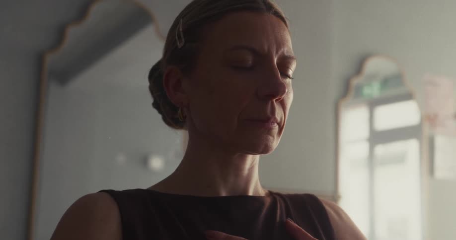 A serene woman practices mindful meditation with her eyes closed in a peaceful, sunlit room, shown in a slow-motion close-up