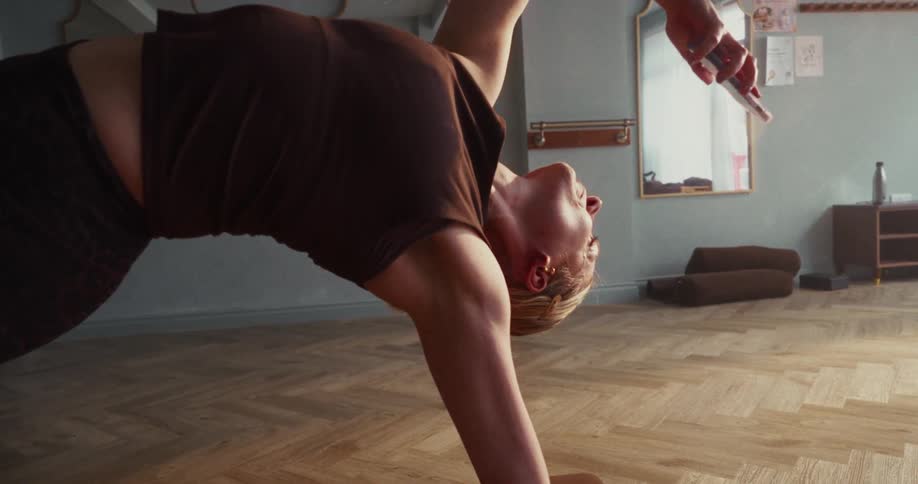 A flexible woman holds the Wild Thing yoga pose while taking a selfie on her phone in a sunlit studio