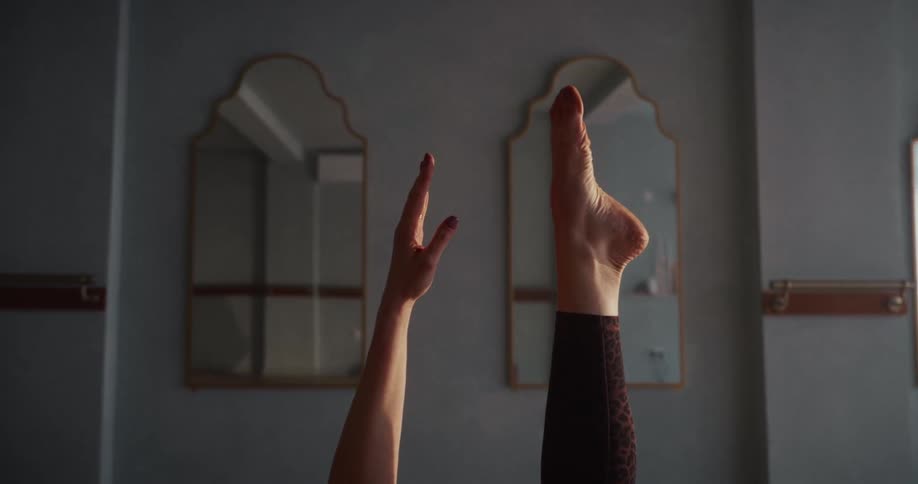 A woman's hand and foot are held steady in a yoga pose in a serene, sunlit room