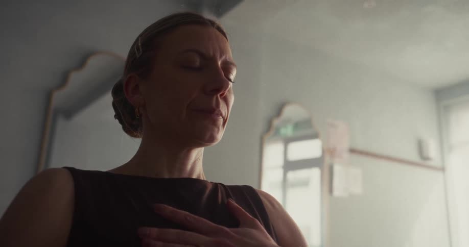 A woman with her eyes closed meditates peacefully with her hand on her heart in a softly lit room