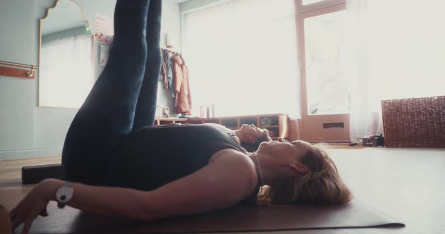 A woman gracefully transitions from a plow pose to a bridge pose in slow motion during a sunlit yoga class