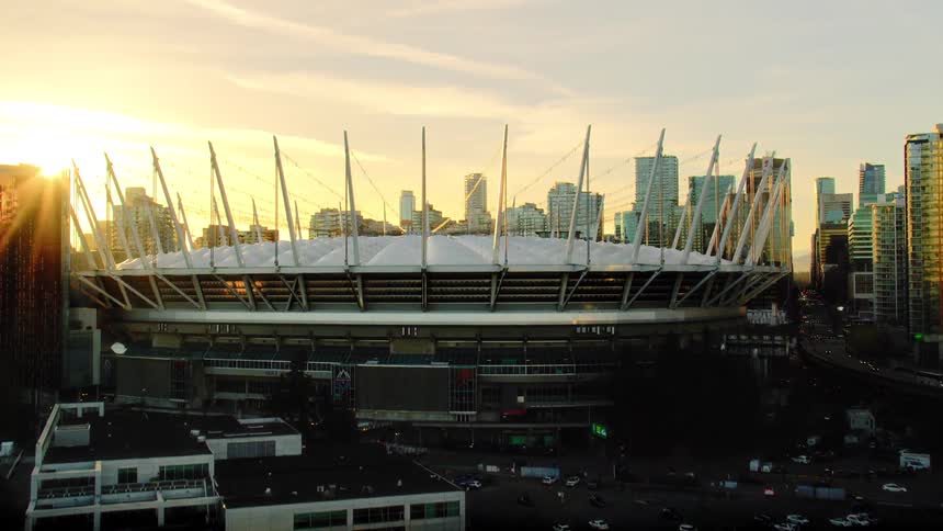 A dramatic aerial shot tilts up from the water to reveal the iconic BC Place stadium against the Vancouver city skyline during a golden sunset